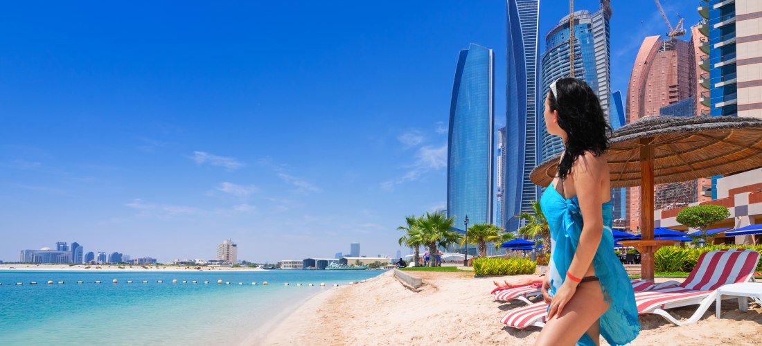 Frau in blauem Strandkleid steht am Sandstrand in Abu Dhabi mit Blick auf das türkisfarbene Wasser und die modernen Wolkenkratzer der Skyline