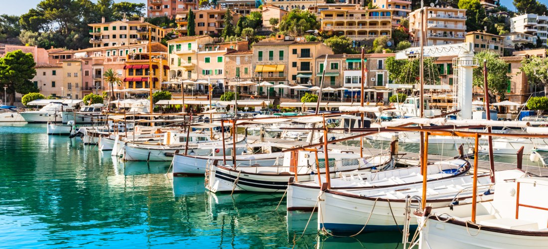 Traditionelle Holzboote im Hafen von Port de Sóller auf Mallorca, vor farbenfrohen Gebäuden und grünen Hügeln
