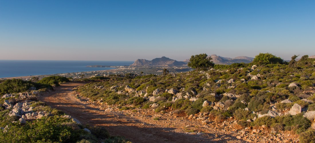 Staubiger Wanderpfad mit Blick auf eine grüne, felsige Hügellandschaft und die weite Küste von Kos. Im Hintergrund liegen Bergketten, die von der untergehenden Sonne in warmes Licht getaucht werden.