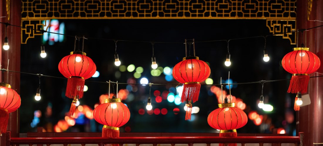 Das Bild zeigt eine nächtliche Szene mit traditionellen roten chinesischen Laternen, die an schwarzen Kabeln zusammen mit kleinen Glühbirnen aufgehängt sind. Sie hängen vor einer verzierten Balustrade aus rotem Holz mit goldenen Mustern. Im Hintergrund le