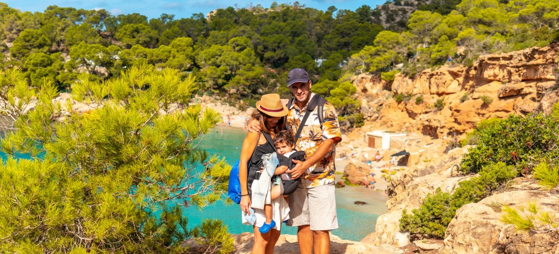 Eine junge Familie steht an einem Aussichtspunkt über einer türkisblauen Bucht auf Ibiza. Die Eltern halten ein Kleinkind im Arm, alle lächeln in die Kamera. Im Hintergrund sind Felsen, grüne Pinien und das klare Wasser zu sehen. Die Szene vermittelt Urla