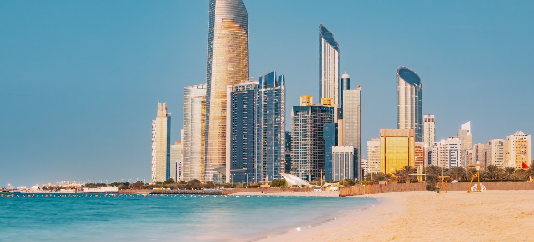 Sonnenuntergang über der Skyline von Abu Dhabi mit goldfarbenen Hochhäusern, türkisblauem Meer und einem Sandstrand im Vordergrund