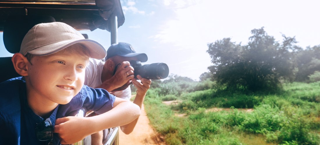 Ein Junge mit heller Kappe lehnt sich aus einem Safarifahrzeug und schaut neugierig in die Landschaft. Hinter ihm fotografiert ein Mann mit einer Kamera. Das Fahrzeug fährt auf einem sandigen Weg durch grünes Buschland unter leicht bewölktem Himmel. Die S