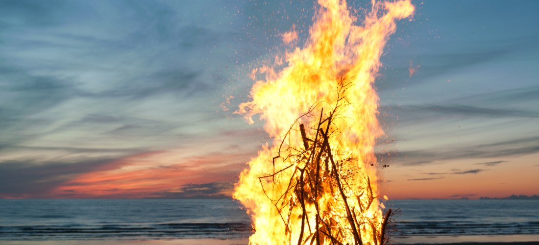 Großes Lagerfeuer aus aufgeschichteten Ästen brennt am Strand bei Sonnenuntergang. Der Himmel ist blau-orange gefärbt, das Meer liegt ruhig im Hintergrund.