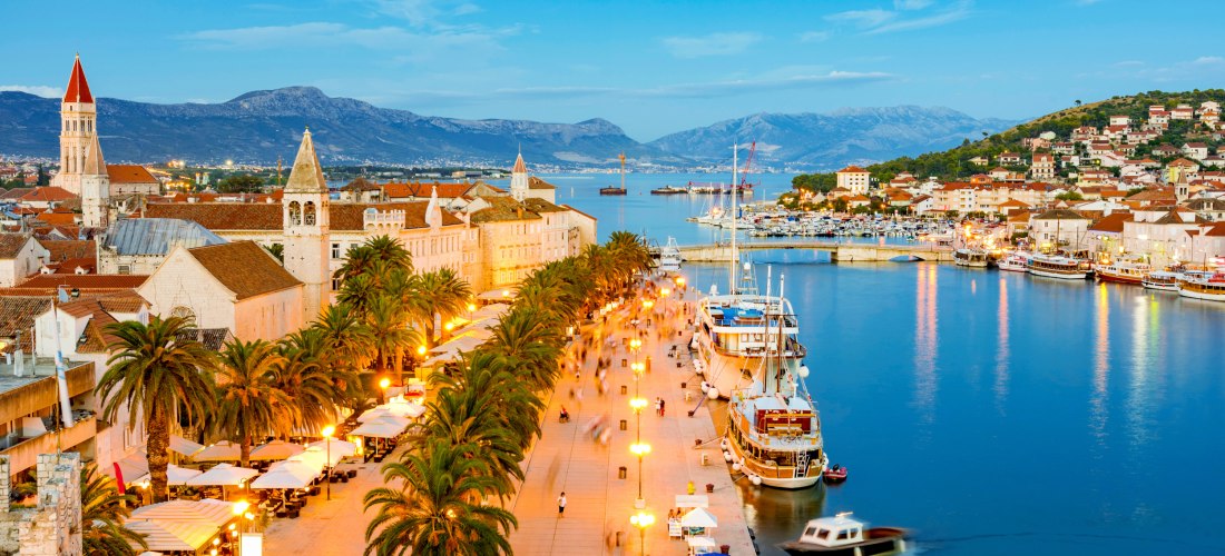 Abendstimmung an der Uferpromenade von Trogir in Kroatien. Entlang der beleuchteten Palmenallee flanieren Menschen, während Segelboote und Yachten im Hafen liegen. Im Hintergrund leuchten die historischen Gebäude und Kirchtürme der Altstadt, umrahmt von d