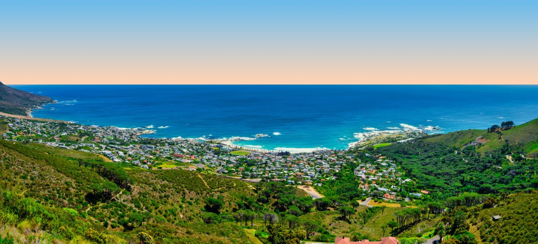 Weitläufiges Küstenpanorama in Südafrika mit Blick auf eine Stadt am Meer, umgeben von grünen Hügeln. Im Hintergrund trifft der tiefblaue Atlantik auf den Horizont bei orangem Sonnenuntergang.
