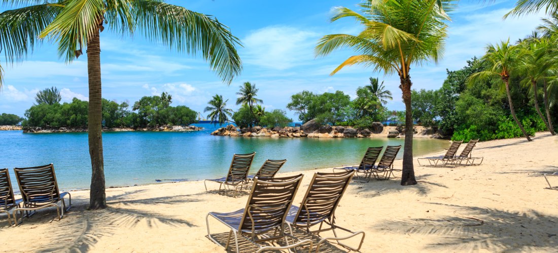 Mehrere leere Liegestühle stehen unter Palmen an einem ruhigen Sandstrand in Singapur. Im Hintergrund liegen kleine grüne Inseln in einer türkisblauen Bucht unter blauem Himmel mit weißen Wolken
