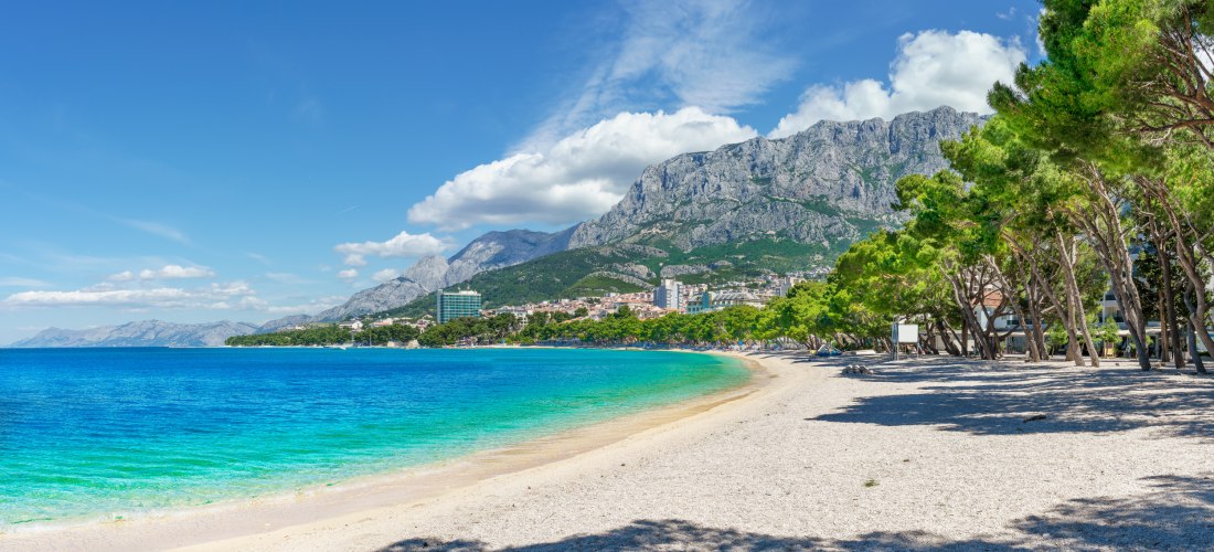 Langer Kiesstrand mit türkisblauem Wasser, im Hintergrund eine Stadt vor imposanten, grauen Gebirgszügen. Am rechten Bildrand spenden Pinienbäume Schatten auf den Strand.