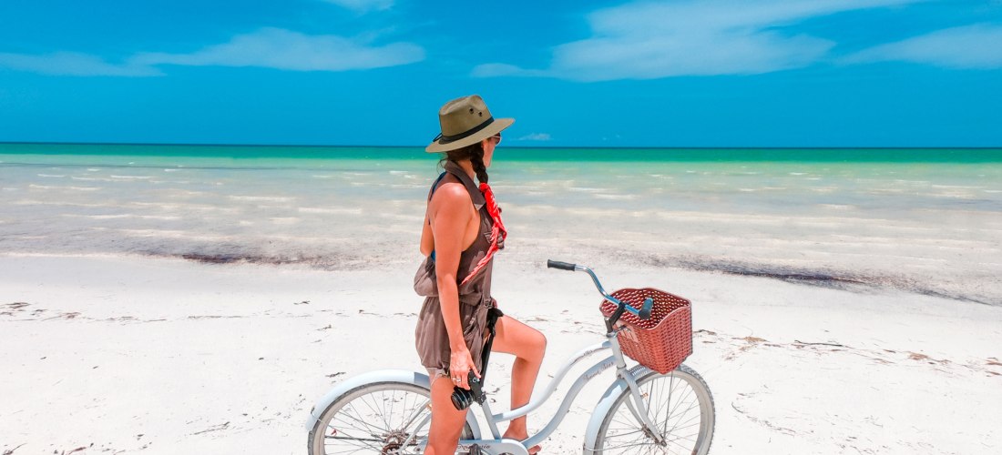 Junge Frau mit Fahrrad am weißen Sandstrand der Isla Holbox in Mexiko, türkisfarbenes Meer im Hintergrund – entspannter Karibikurlaub 2025/2026
