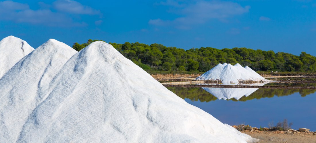 Salzberge in einer Saline auf Mallorca. Die Salzberge sind hoch und weiß, sehen aus wie kleine schneebedeckte Hügel, die sich vor einem klaren blauen Himmel und einem grünen Kiefernwald abheben. Im Vordergrund spiegelt sich der Himmel und die Salzberge in