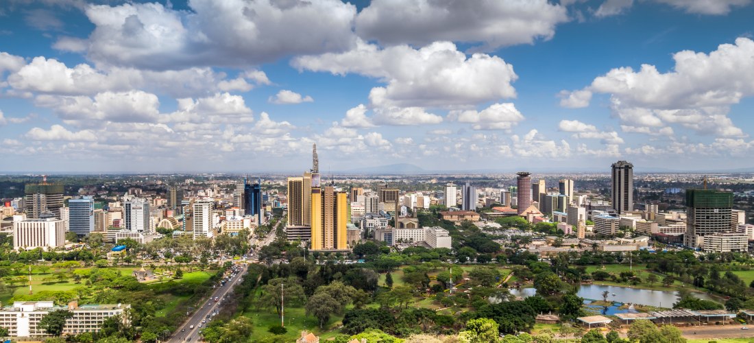 Panoramablick über Nairobi mit moderner Skyline, Hochhäusern, Grünflächen und Straßenverkehr. Weiße Wolken schweben über der Stadt, im Vordergrund ein Park mit Bäumen und einer Kirche