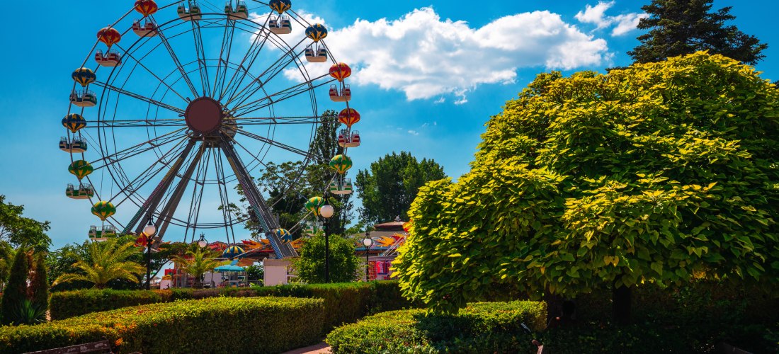 Großes Riesenrad in einem Freizeitpark am Meer, umgeben von gepflegten Gärten, Bäumen und Parkbänken. Der Himmel ist blau mit einigen weißen Wolken