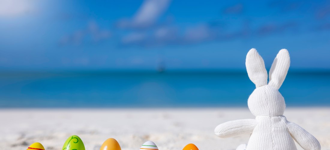 Ein gestrickter weißer Stoffhase sitzt im Sand am Strand und blickt aufs Meer. Vor ihm liegen bunte Ostereier im Sand, dahinter der türkisfarbene Ozean und blauer Himmel.