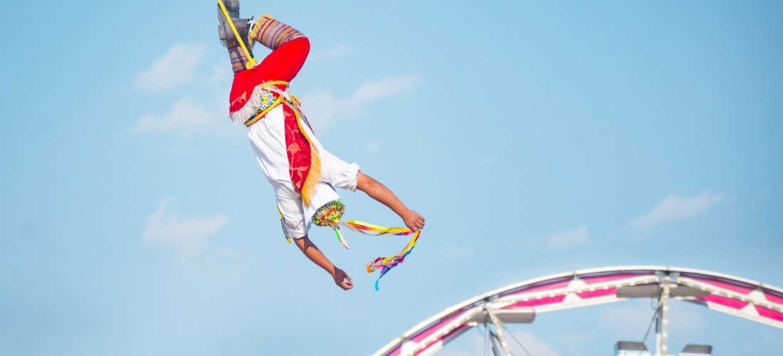 Ein Volador de Papantla, ein traditioneller mexikanischer Lufttänzer, schwebt kopfüber an einem Seil, gekleidet in ein buntes Kostüm mit roten Hosen, weißen Oberteilen und bunten Bändern, mit einem Riesenrad im Hintergrund