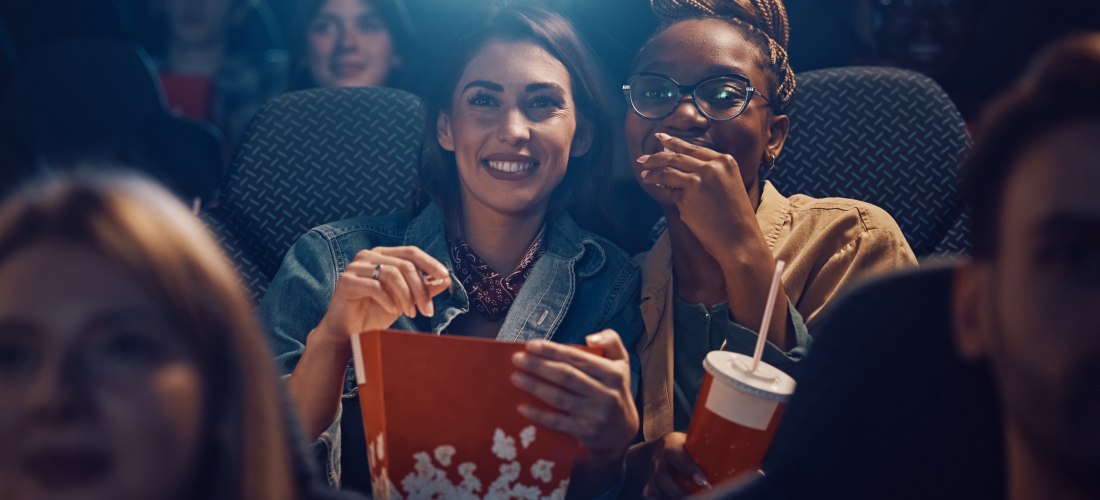 Zwei Frauen sitzen lachend im Kino, die vordere hält eine große Box Popcorn, die andere einen Softdrink. Sie schauen gespannt nach vorn, während im Hintergrund weitere Zuschauer zu sehen sind