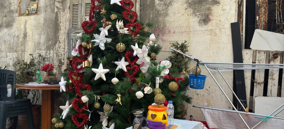 Ein liebevoll geschmückter Weihnachtsbaum mit roten Herzen, weißen Sternen und goldenen Kugeln steht vor einer verwitterten Hauswand in einer griechischen Gasse. Daneben ein kleiner Tisch mit einer Decke, Geschirr und Haushaltsgegenständen, sowie ein Wäsc