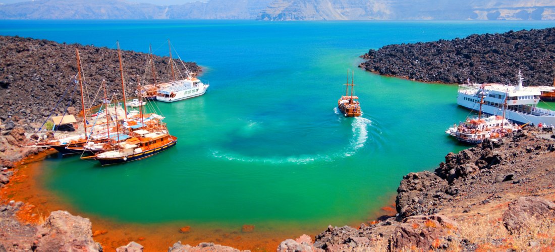 Nea Kameni: Ausflugsboote liegen in einer leuchtend grün-türkisen Thermalbucht, schwarze Lavafelsen und Caldera-Kulisse im Hintergrund