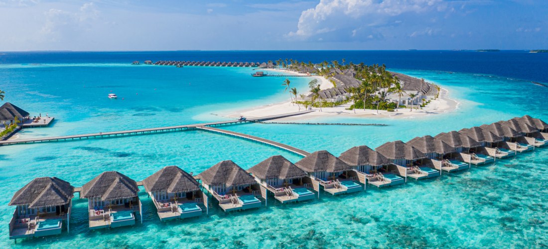 Luftaufnahme einer tropischen Insel mit weißen Sandstränden, Palmen und zahlreichen Wasserbungalows mit Strohdächern, die in Reihen über türkisblauem, kristallklarem Meer angeordnet sind. Die Anlage ist über Holzstege verbunden. Im Hintergrund tiefblaues 