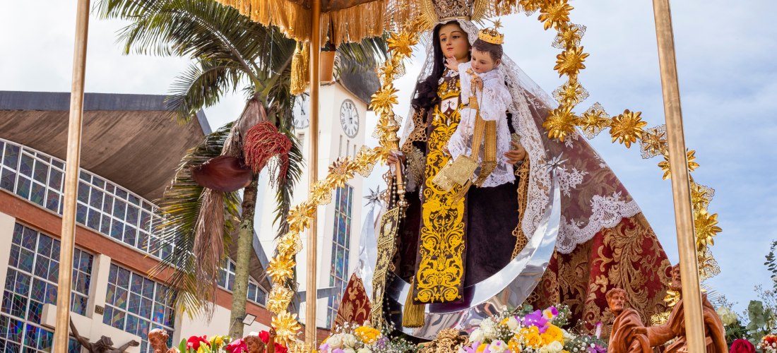 Religiöse Prozession mit kunstvoll geschmückter Marienstatue, die das Jesuskind trägt, unter einem Baldachin vor einer Kirche mit Palmen im Hintergrund