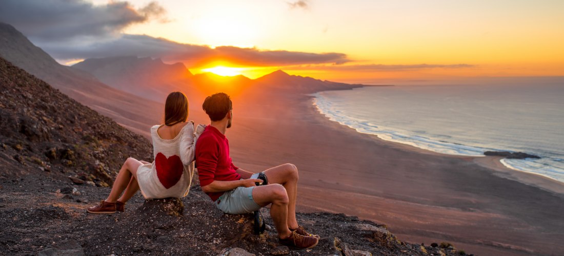 Ein Paar sitzt auf einem Felsen und schaut in den Sonnenuntergang über einer weiten Küstenlandschaft mit Bergen und Meer. Die Frau trägt einen Pullover mit rotem Herz auf dem Rücken