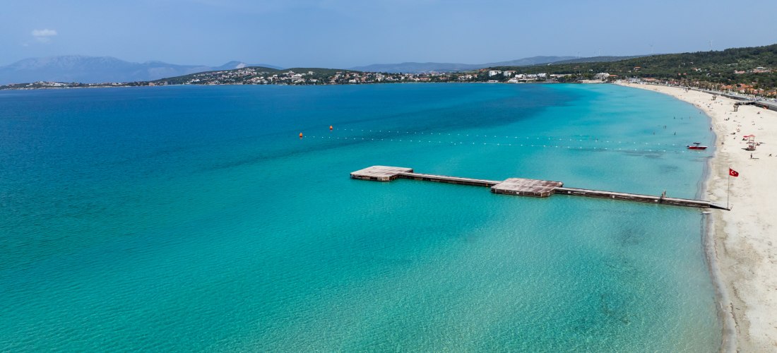 Panoramaaufnahme eines langen Sandstrands mit kristallklarem, türkisfarbenem Wasser an der türkischen Ägäis. Ein hölzerner Badesteg ragt ins Meer, im Hintergrund sind sanfte Hügel und Küstenorte zu sehen.