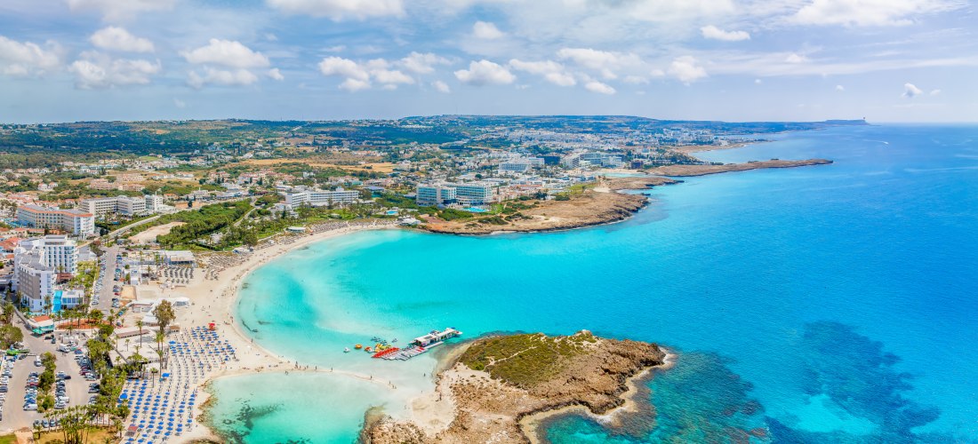 Panoramablick auf Ayia Napa mit weißem Sandstrand, türkisfarbenem Meer und zahlreichen Hotels entlang der Küste.