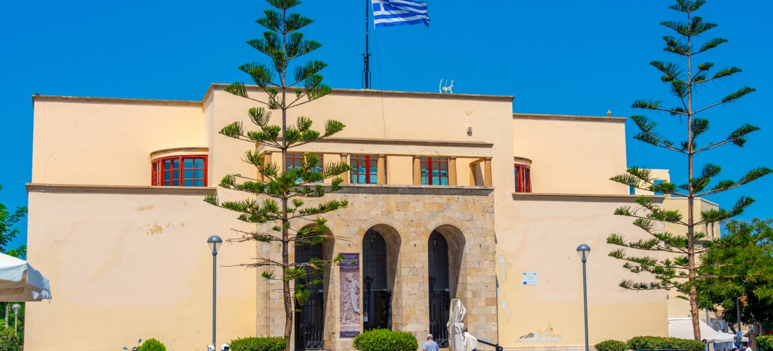 Symmetrisches Museumsgebäude mit drei Rundbögen und griechischer Flagge auf dem Dach, davor ein Platz mit Bäumen und Menschen bei sonnigem Wetter