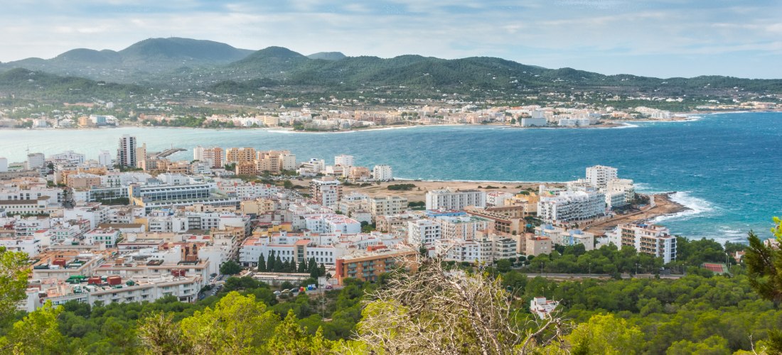 Blick auf die Küstenstadt Sant Antoni de Portmany auf Ibiza. Im Vordergrund grüne Pinienwälder, dahinter zahlreiche weiße und bunte Häuser bis hin zum Meer. Im Hintergrund die hügelige Landschaft der Insel.