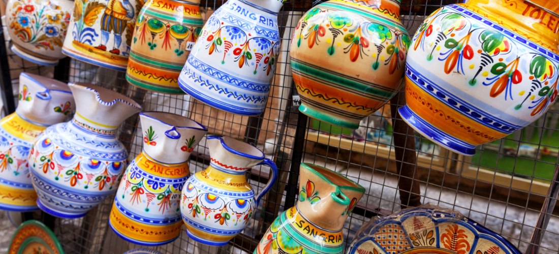 Bunte handbemalte Tonkrüge hängen an einem Gitter. Die Krüge sind mit floralen Mustern, Schriftzügen wie 'Sangria' und traditionellen spanischen Motiven verziert.