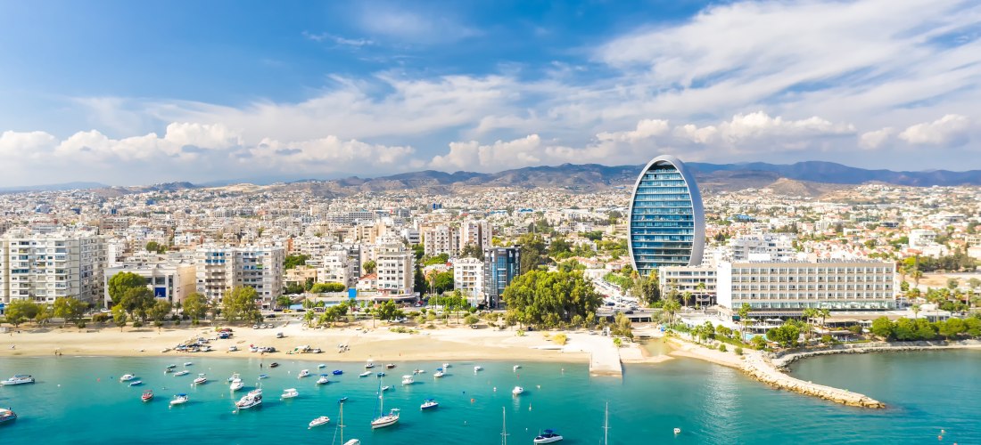 Panoramablick auf Limassol mit Strand, Segelbooten und dem markanten Oval Tower an der Küste.