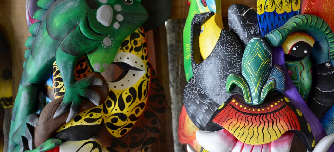 Bunte, handgefertigte Holzmasken mit Tiermotiven, darunter ein grünes Leguan, ein Tukan und weitere fantasievolle, farbenfrohe Designs, ausgestellt an einer Holzwand