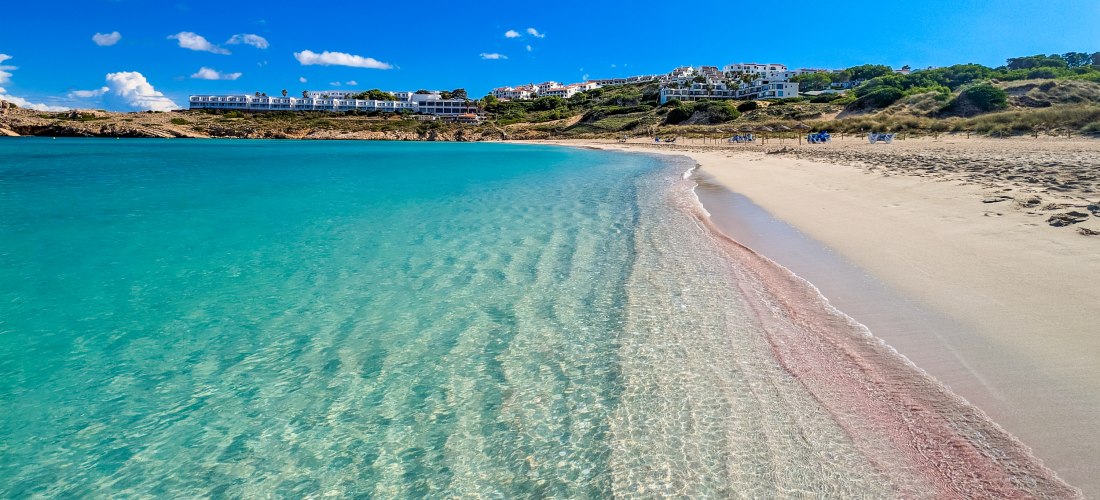 Flacher Sandstrand mit kristallklarem Wasser auf Menorca. Das Meer schimmert in türkis und verläuft sanft in den beigen Sand. Eine rötliche Algenlinie trennt den feuchten vom trockenen Sand. Im Hintergrund sind weiße Häuser und grüne Hügel zu sehen unter 