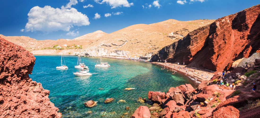 Blick auf den berühmten Red Beach auf Santorini, eingerahmt von markanten roten Felsformationen. Türkisblaues Wasser mit mehreren Segelbooten, die in der kleinen Bucht vor Anker liegen. Im Hintergrund trockene, ockerfarbene Hügel unter einem strahlend bla