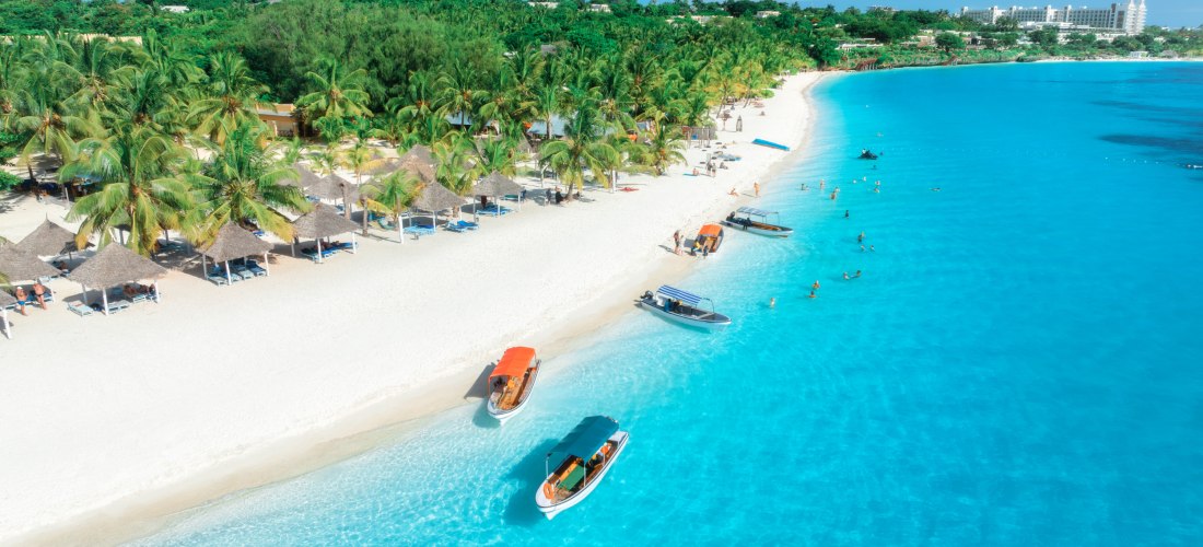Traumstrand auf Sansibar mit Palmen, türkisfarbenem Wasser, Booten und entspannenden Urlaubern am weißen Sand.