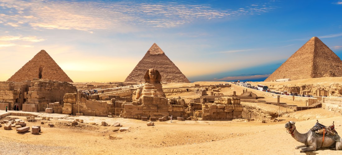 Panoramablick auf die Pyramiden von Gizeh in Ägypten bei Sonnenuntergang. In der Mitte die berühmte Sphinx, im Vordergrund ein liegendes Kamel mit Sattel. Touristen sind auf dem Gelände unterwegs, der Himmel ist goldblau gefärbt.