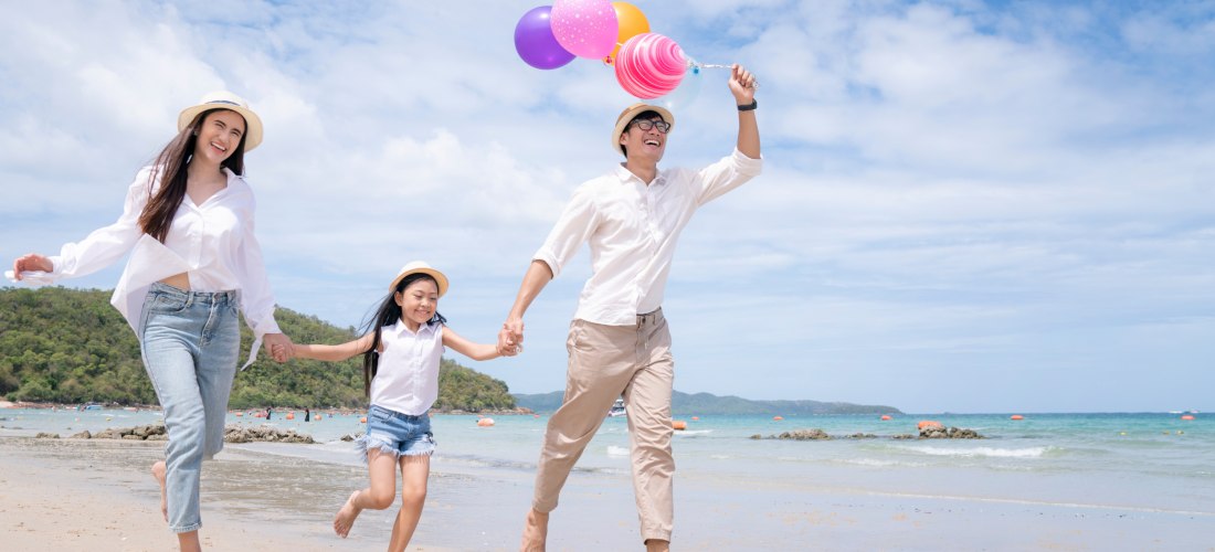 Lachende Familie mit Kind läuft barfuß am Strand entlang. Der Vater hält bunte Luftballons, alle tragen helle Kleidung und Hüte. Im Hintergrund türkisblaues Meer und grüne Hügel