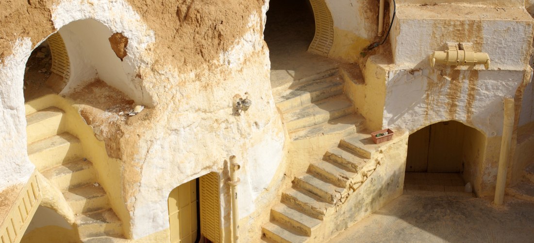 Höhlenwohnungen in Matmata, Tunesien. In den ockerfarbenen Felsen sind weiß gestrichene Türen und Fenster eingelassen. Kleine, verwinkelte Treppen führen zu den Eingängen, die in den Fels gehauen sind. Die Szene vermittelt ein Gefühl von Geschichte und tr