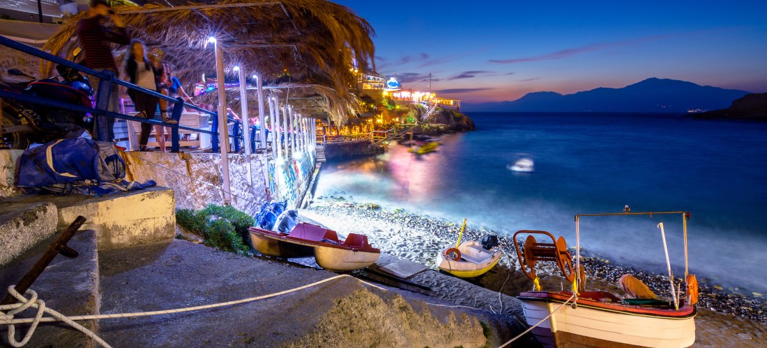 Ein kleiner Küstenort auf Kreta bei Abenddämmerung. Am Ufer liegen bunte Fischerboote, die Wellen schlagen sanft gegen die Steinmauer. Entlang der Promenade sitzen Menschen unter strohgedeckten Dächern in beleuchteten Tavernen. Der Himmel ist dunkelblau, 