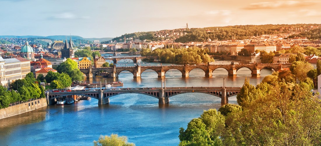 Panorama von Prag mit Moldau und mehreren Brücken, darunter die Karlsbrücke; Altstadtdächer, Türme und viel Grün im warmen Licht.