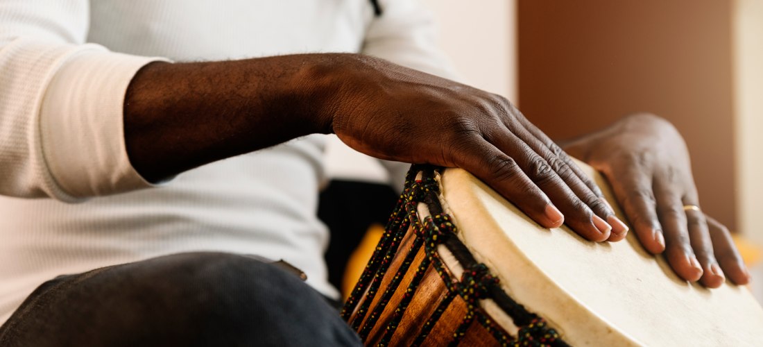 Ein Mann spielt eine Djembe, eine traditionelle Trommel. Die Aufnahme zeigt den Oberkörper und die Hände, wie sie auf das Trommelfell schlagen. Der Mann trägt ein weißes Langarmshirt und sitzt mit überkreuzten Beinen. Die Djembe ist mit bunten Seilen verz