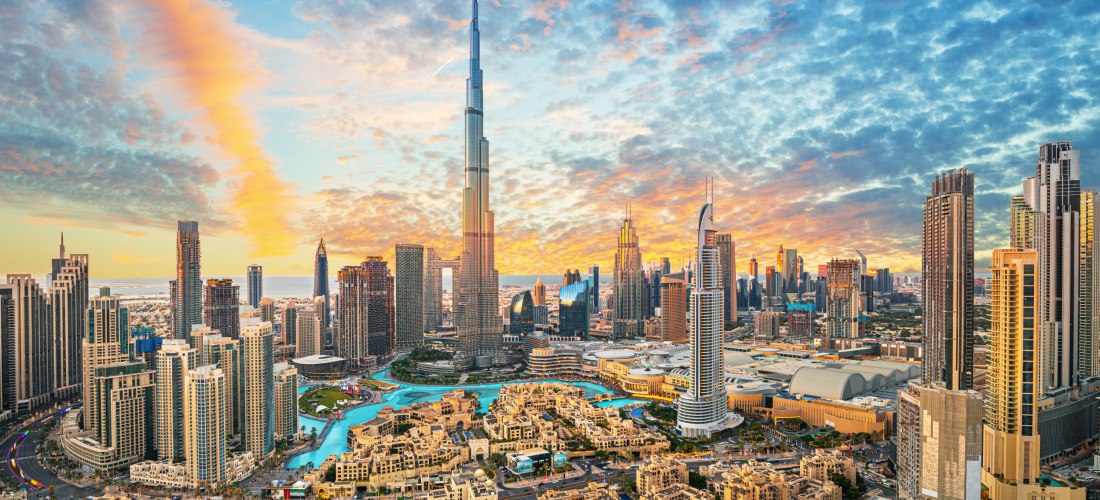 Dubai - Downtown Panorama der Skyline von Downtown Dubai bei Sonnenuntergang. Im Zentrum steht der Burj Khalifa, umgeben von modernen Hochhäusern, Wasserläufen und Hotels. Der Himmel zeigt eine dramatische Wolkenformation mit orange-blauen Lichtverläufen. Im Vordergrund s