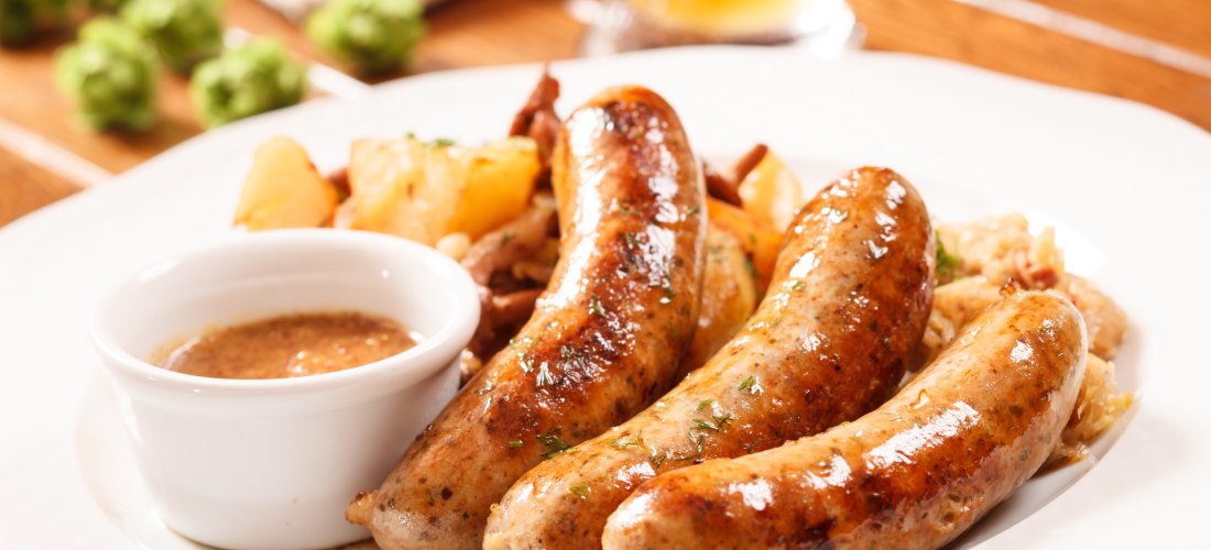 Drei gebratene Bratwürste auf einem weißen Teller, serviert mit gebratenen Kartoffeln, Sauerkraut und Senf in einer kleinen weißen Schale. Im Hintergrund unscharf ein Glas Bier und grüne Dekoration.