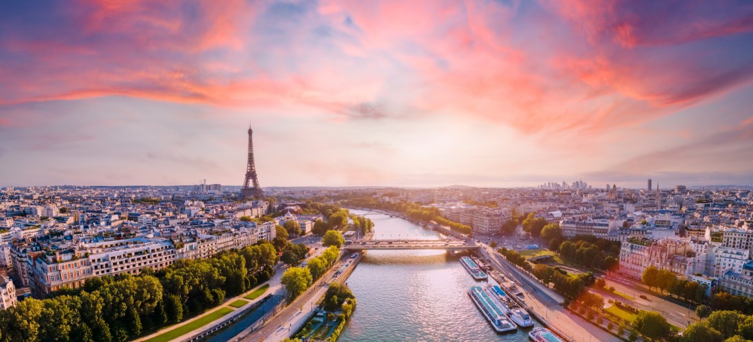 Weitwinkel-Panorama von Paris bei Sonnenuntergang: die Seine durchschneidet die Stadt, in der Mitte der Eiffelturm; Uferpromenaden, Brücken und Hausdächer unter rosa Wolken