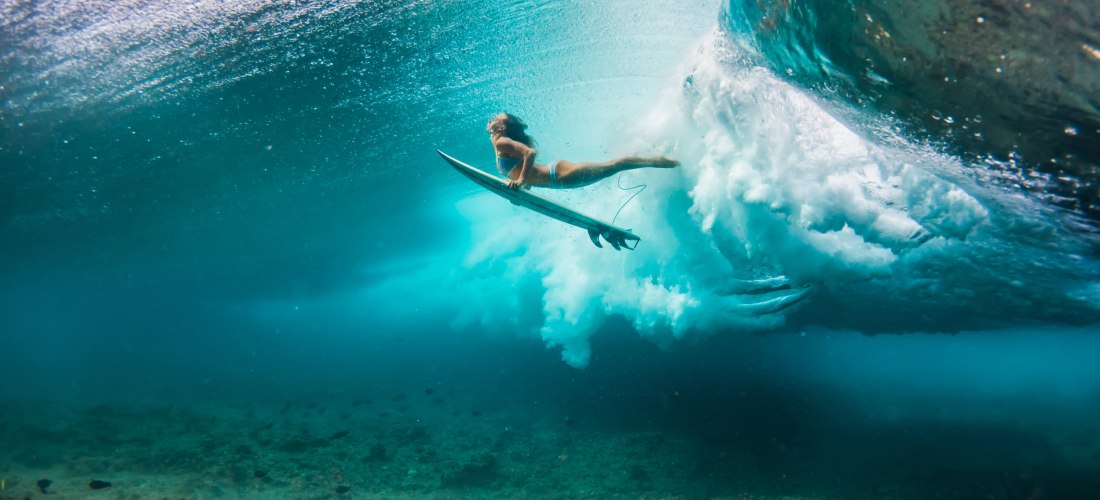 Eine Surferin gleitet mit ihrem Board unter einer gewaltigen Welle hindurch. Die Unterwasserperspektive              zeigt die kraftvolle Dynamik des Meeres und die faszinierende Welt unter der Oberfläche.