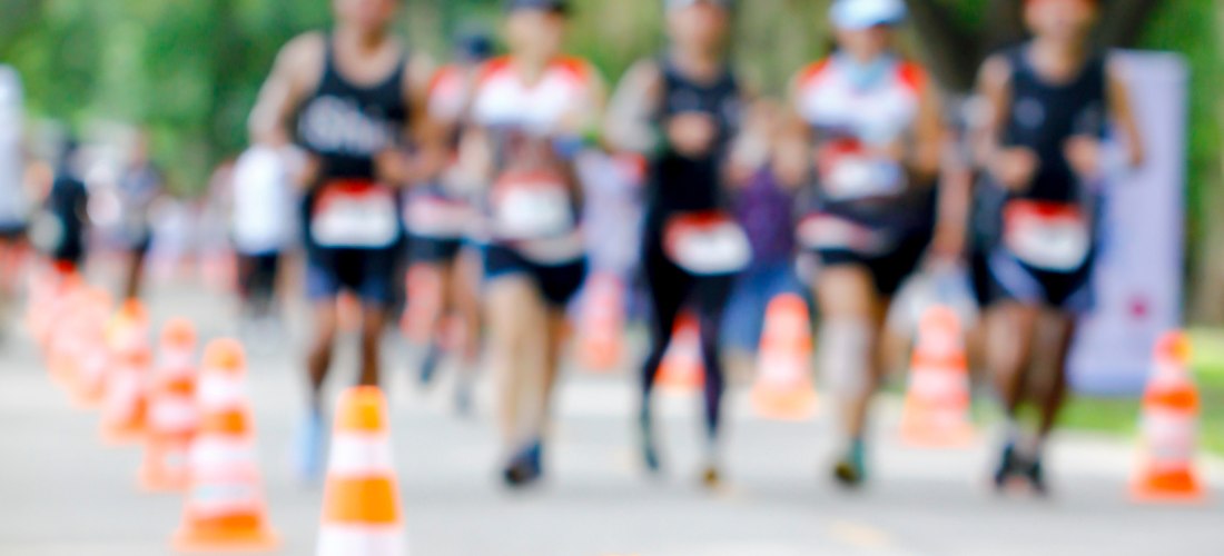 Laufveranstaltung auf einer von Bäumen gesäumten Straße, im Vordergrund orange-weiße Verkehrskegel, im Hintergrund Läuferinnen und Läufer in Sportkleidung, teils unscharf.