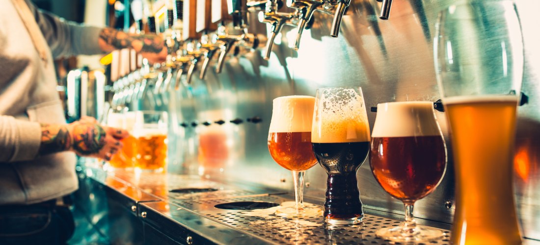 Zapfanlage in einer Bar: Mehrere Biersorten werden frisch gezapft. Im Vordergrund stehen verschiedene Biergläser gefüllt mit Bierschaum. Ein Barkeeper mit tätowiertem Arm zapft Bier
