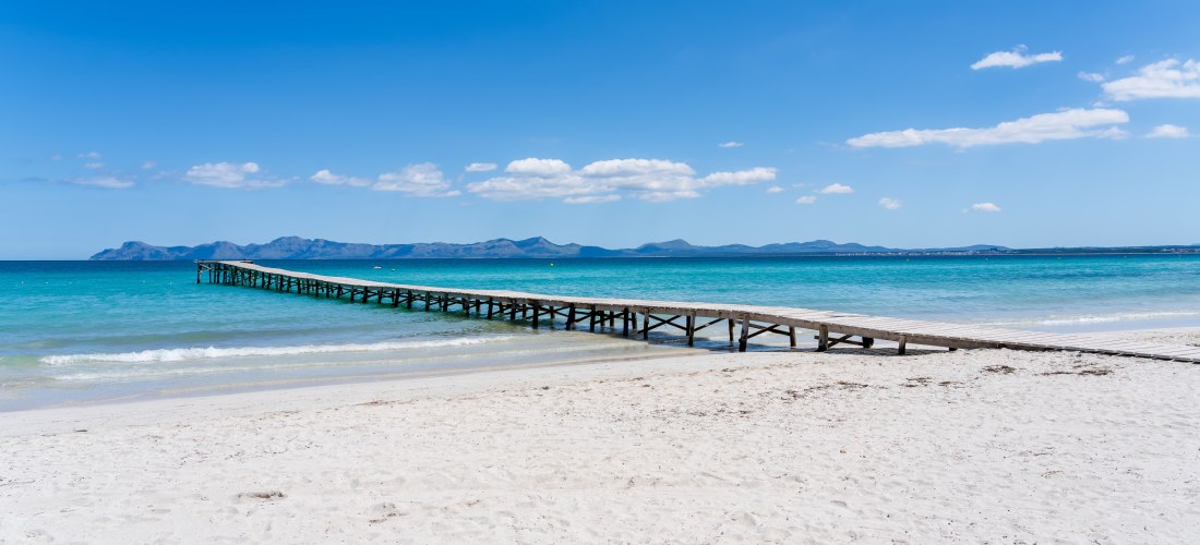 Holzsteg, der vom weißen Sandstrand weit ins türkisfarbene Meer hinausragt, im Hintergrund eine Gebirgskette unter blauem Himmel mit wenigen Wolken.