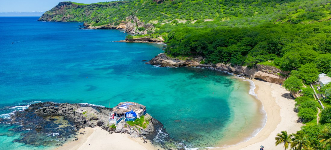 Luftaufnahme einer traumhaften Bucht auf den Kapverden mit grün bewaldeten Hügeln, steilen Felsen und klarem, türkisfarbenem Wasser. In der Mitte eine kleine Felseninsel mit einer Strandbar. Der helle Sandstrand ist von Palmen gesäumt.