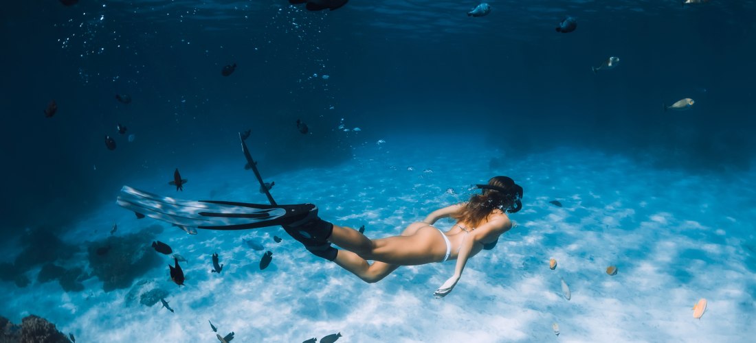 Eine Frau taucht mit Schnorchel und Flossen durch klares, türkisfarbenes Wasser über einen sandigen Meeresboden. Um sie herum schwimmen zahlreiche kleine bunte Fische. Die Szene wirkt ruhig und zeigt die Schönheit der Unterwasserwelt