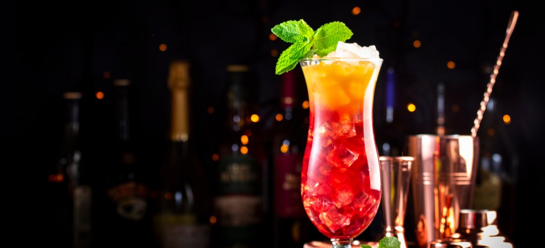 Leuchtend bunter Cocktail in einem hohen Glas, serviert mit Eiswürfeln und Minze. Im Hintergrund stehen Flaschen und Barkeeper-Equipment, umrahmt von stimmungsvollen Lichtern auf dunklem Hintergrund.